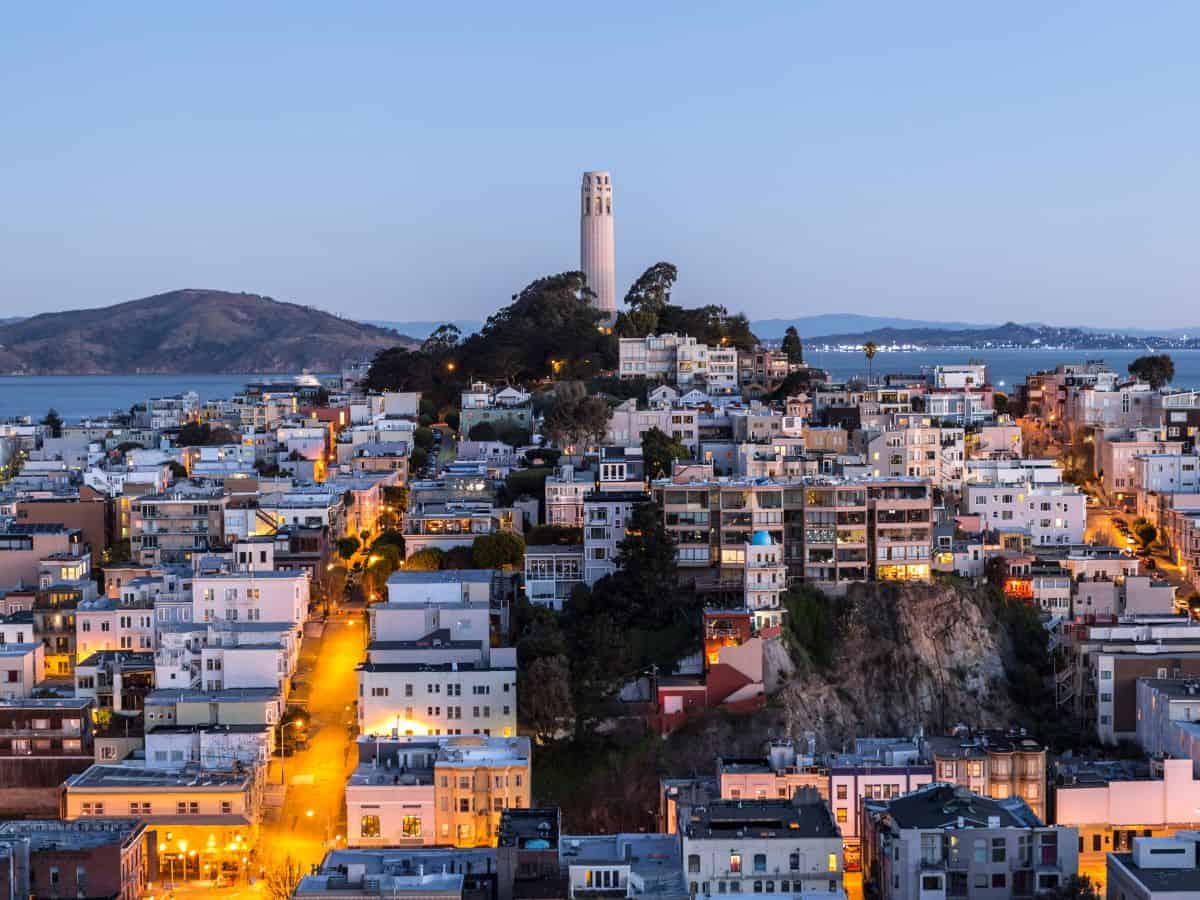 A twilight view of San Francisco's Coit Tower lit up against the evening sky, with the city's densely packed houses stretching out towards the bay. Visiting Coit Tower for its panoramic city views is a popular thing to do in San Francisco.
