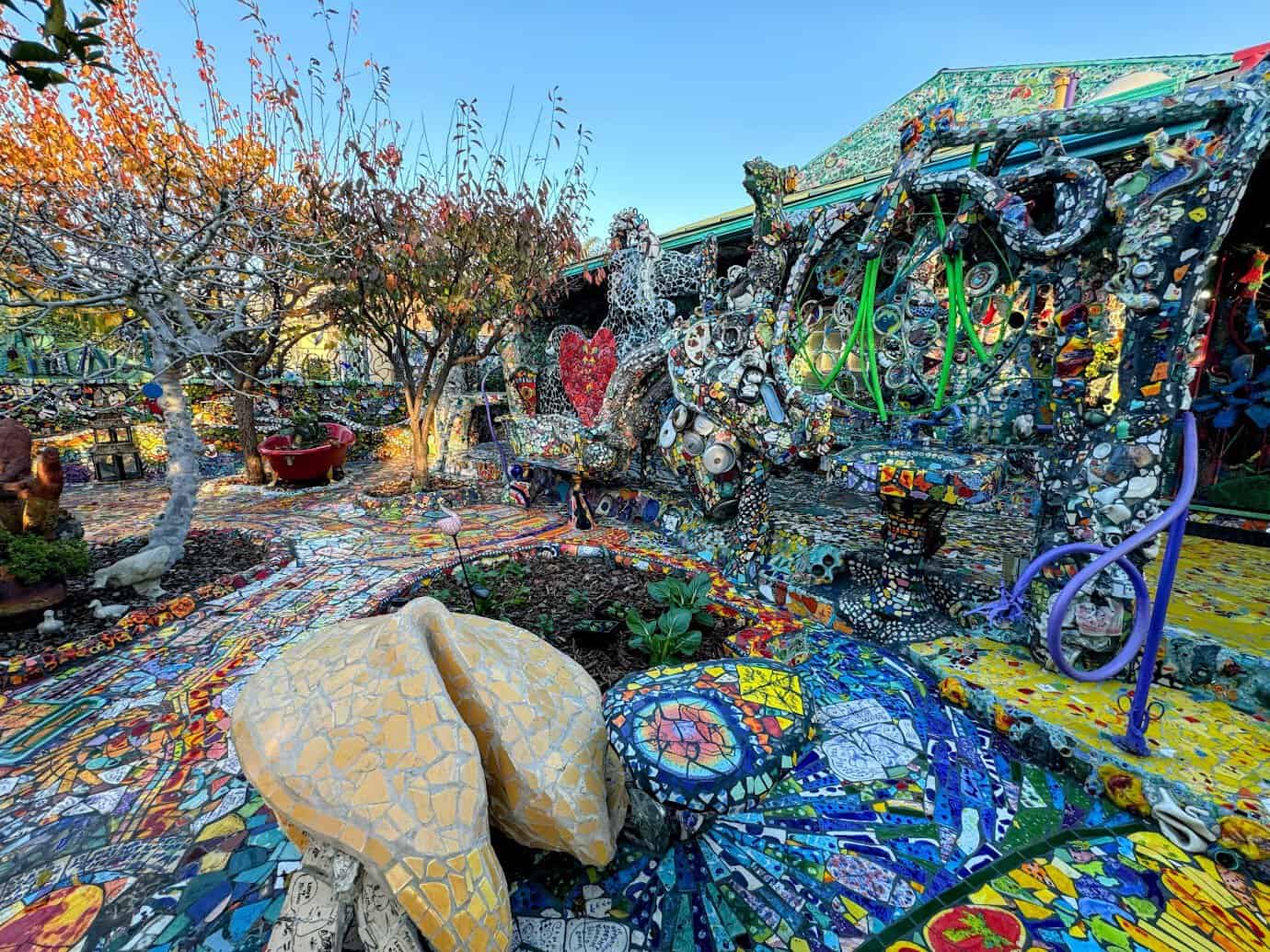An outdoor view of the Mosaic Tile House, showcasing vibrant and intricate mosaic artwork covering nearly every surface. The scene features a colorful walkway made of small, brightly colored tiles leading through the garden. Surrounding the area are creatively decorated structures, including a bench and a table covered in mosaic designs. A large, yellow mosaic sculpture resembling a fortune cookie is prominently displayed in the foreground. Leafless trees with autumn-colored leaves frame the space, while the blue sky provides a striking contrast to the vibrant colors of the artwork. The eclectic and whimsical design creates a lively and artistic atmosphere.