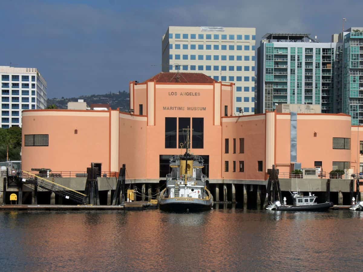 The Los Angeles Maritime Museum building stands by the waterfront with a boat docked in front. The peach-colored structure contrasts with modern high-rise buildings in the background, creating a blend of history and modernity.