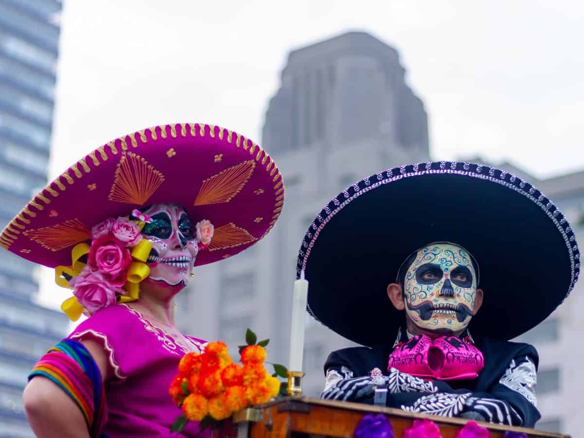 Two individuals dressed in colorful Día de los Muertos costumes and face paint, wearing traditional wide-brimmed hats. Their elaborate outfits celebrate the Mexican holiday, with a cityscape in the background.