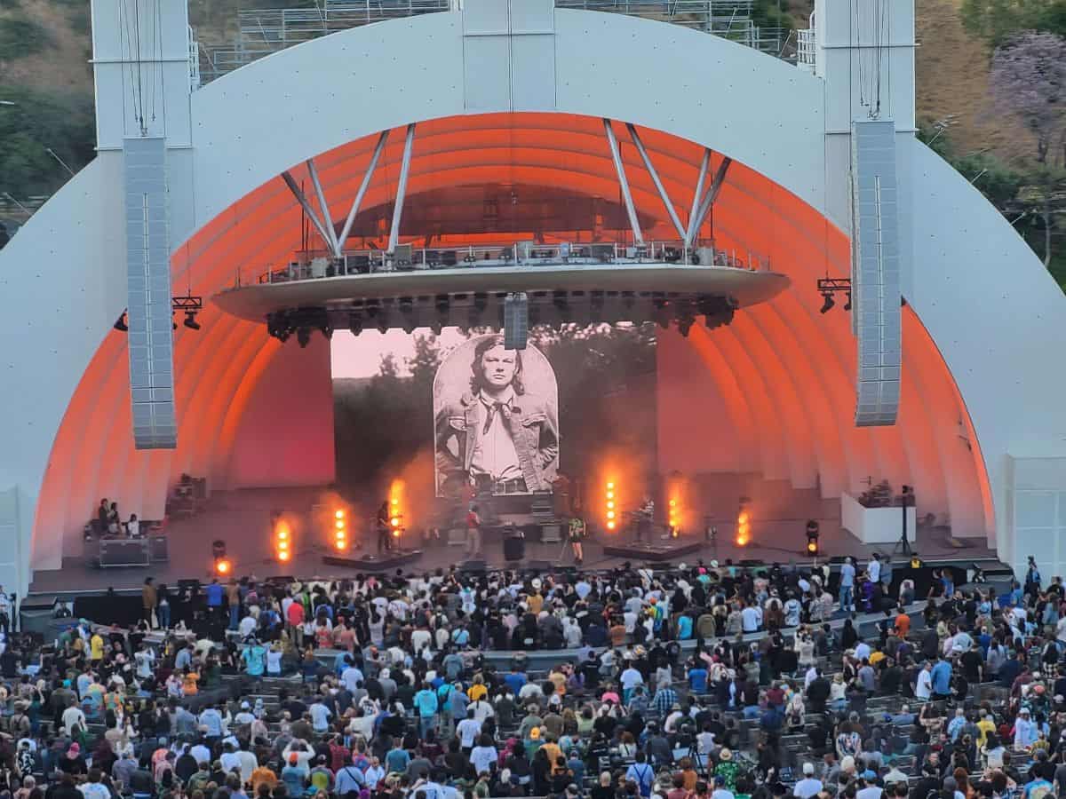 A live performance at the Hollywood Bowl in Los Angeles. The iconic open-air amphitheater features a vibrant stage with lights, a large video screen, and a crowd of concertgoers enjoying the show.