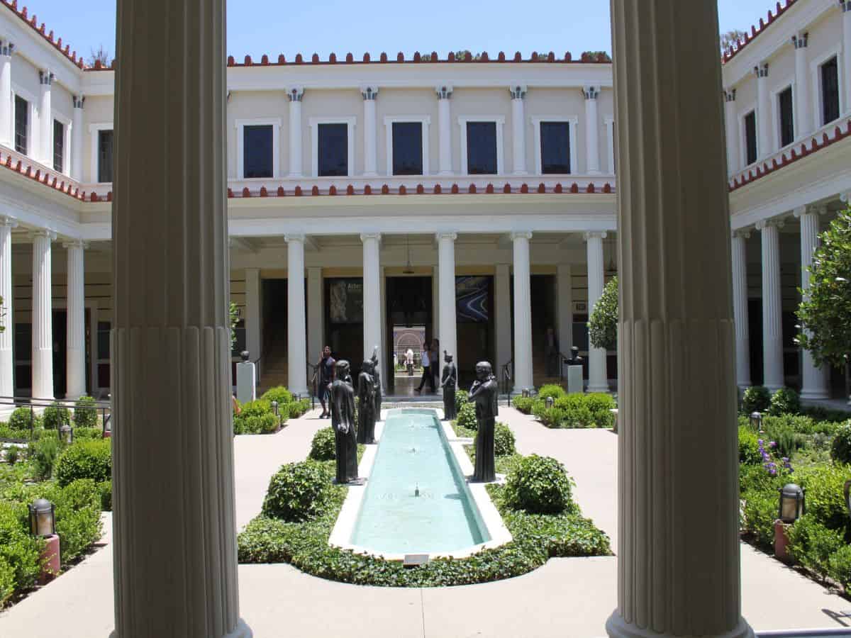 A stunning courtyard at the Getty Villa, showcasing classical Roman-style architecture with tall columns and a central fountain surrounded by greenery. Bronze statues line the fountain, and visitors explore the well-manicured garden in this historic museum in Los Angeles.