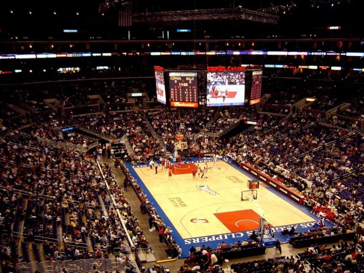 A packed stadium with fans watching a Los Angeles Clippers basketball game at the Staples Center (now Crypto.com Arena). The court is brightly lit, with teams in action and a large scoreboard overhead displaying the game stats.
