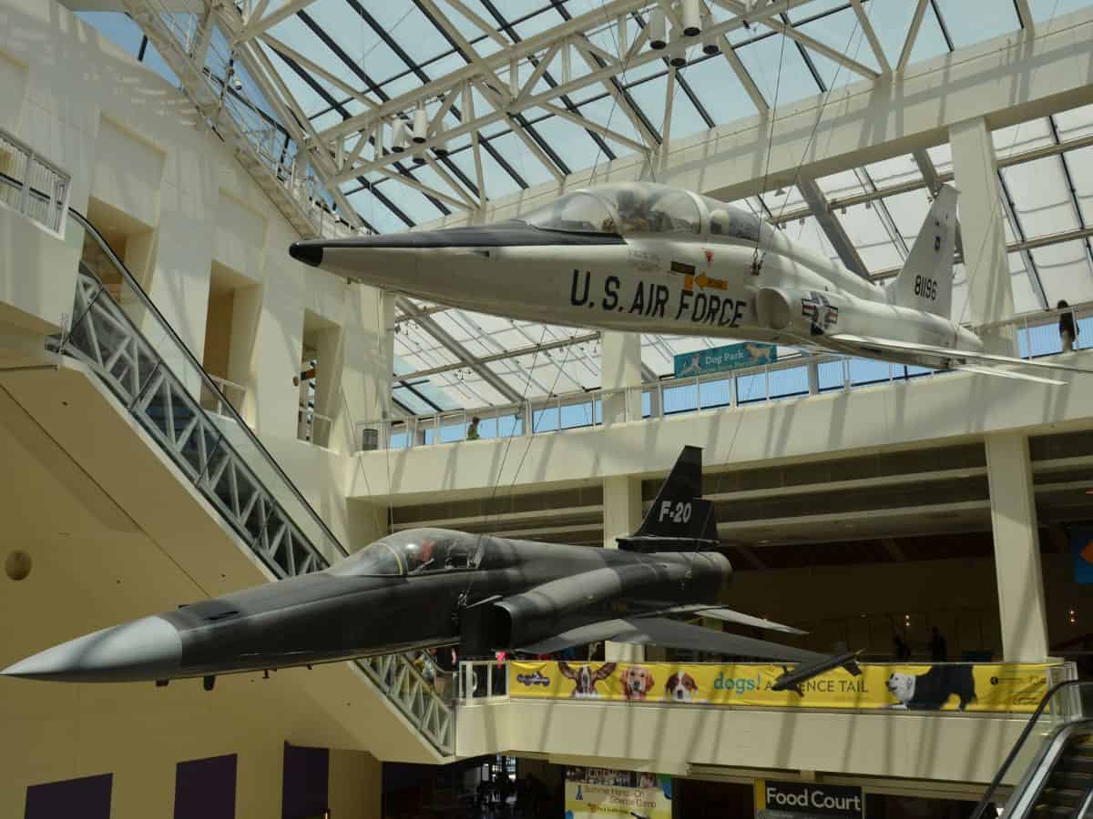 Two suspended fighter jets, one labeled "U.S. Air Force," are on display inside the California Science Center. The planes are showcased under a bright glass ceiling with surrounding exhibits, adding a sense of grandeur to the museum.