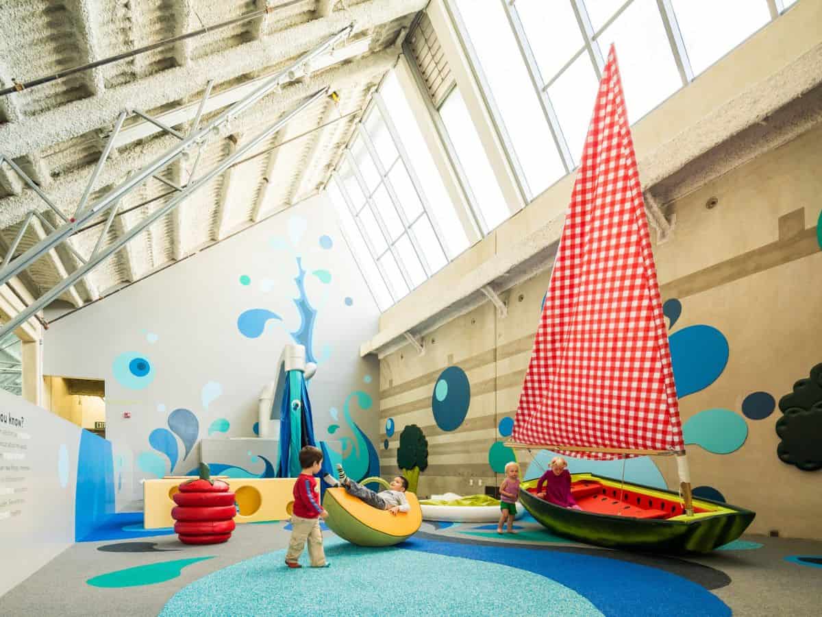 A brightly lit indoor play area at the New Children's Museum with children interacting with colorful, oversized playground structures. One structure resembles a watermelon sailboat with a large red-and-white checkered sail. The play area features soft, safe surfaces, with walls decorated in a playful blue and white theme.