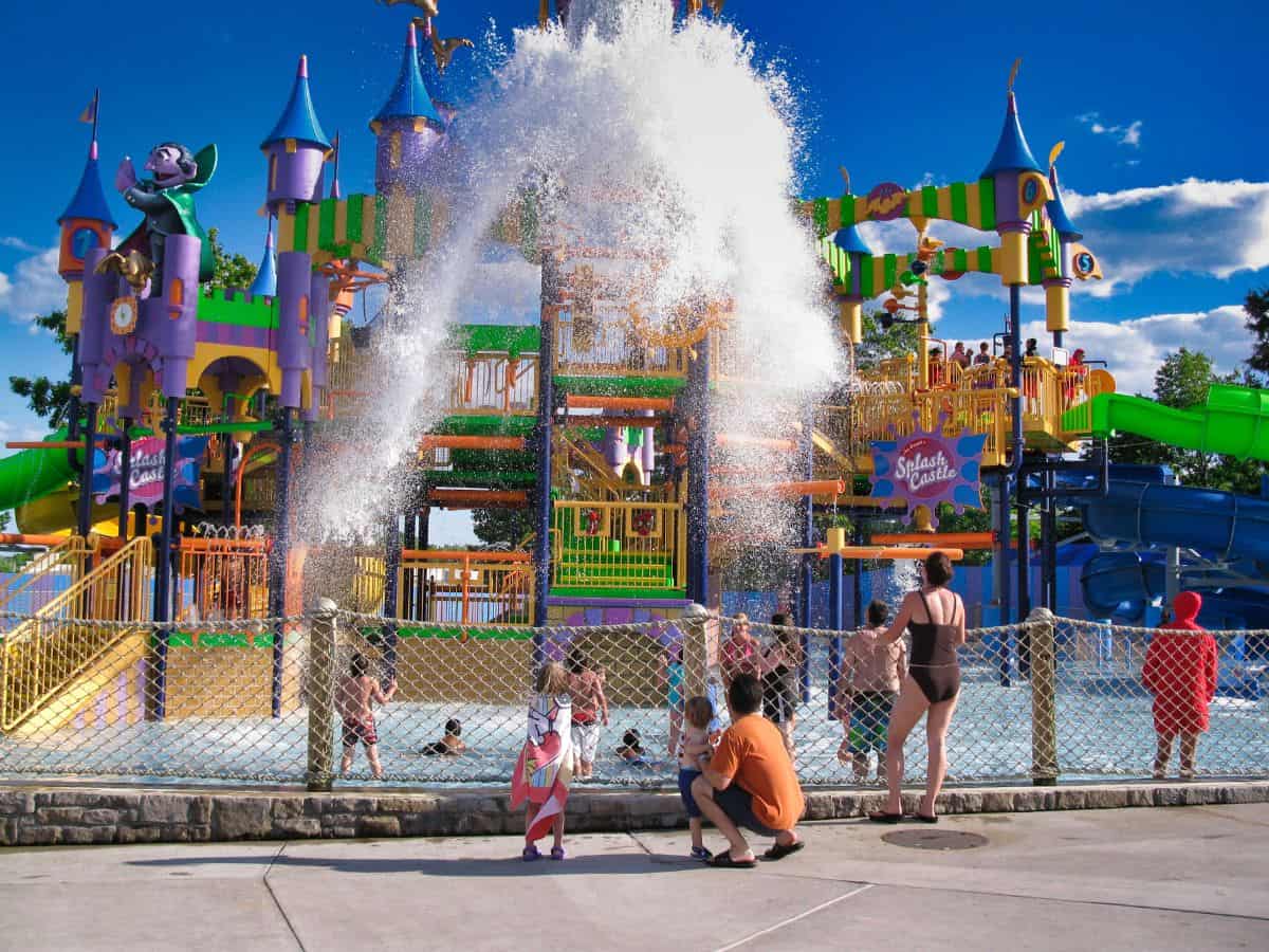 A fun water playground at Sesame Place featuring a colorful castle-like structure with vibrant purple, yellow, and green towers. A huge splash of water is being sprayed from the structure, delighting children and families below. There are multiple water slides and activities surrounding the castle for visitors to enjoy.