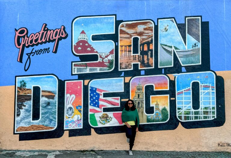 A colorful mural in San Diego reads "Greetings from San Diego" in large block letters. Each letter contains iconic scenes from the city, such as the Hotel del Coronado, a sunset at the pier, a surfer riding waves, and other San Diego landmarks. Shreeyeh stands smiling in front of the mural, wearing sunglasses, a green sweater, and dark pants, casually leaning against the wall. The bright blue sky and vibrant colors of the mural make for a cheerful and welcoming scene.