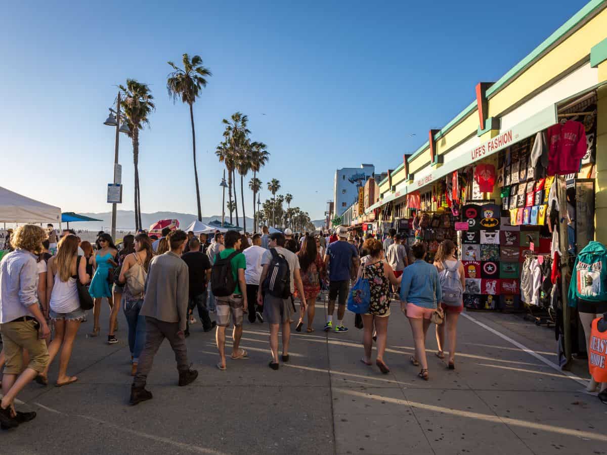 Crowded scene at Venice Beach Boardwalk with diverse groups of people walking, shopping, and enjoying street performances under tall palm trees, a popular thing to do in Los Angeles.