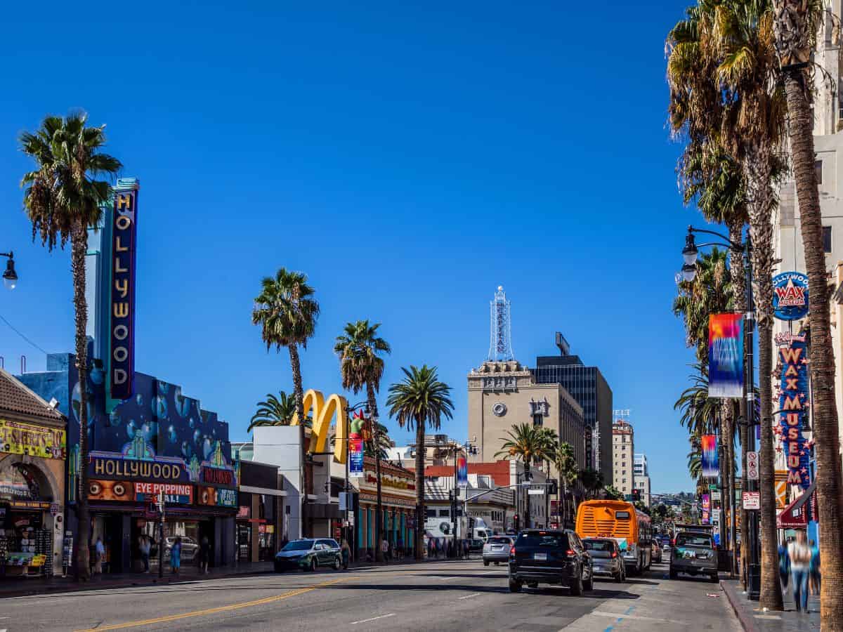 Daytime view of Hollywood Boulevard bustling with activity; visible are the iconic street signs, palm trees, and various entertainment attractions along the sunny street.