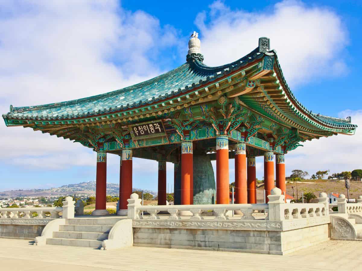 A vivid photo of the Korean Bell of Friendship, a large, ornately decorated green and red pavilion on a clear day, set against a bright blue sky with distant hill views.