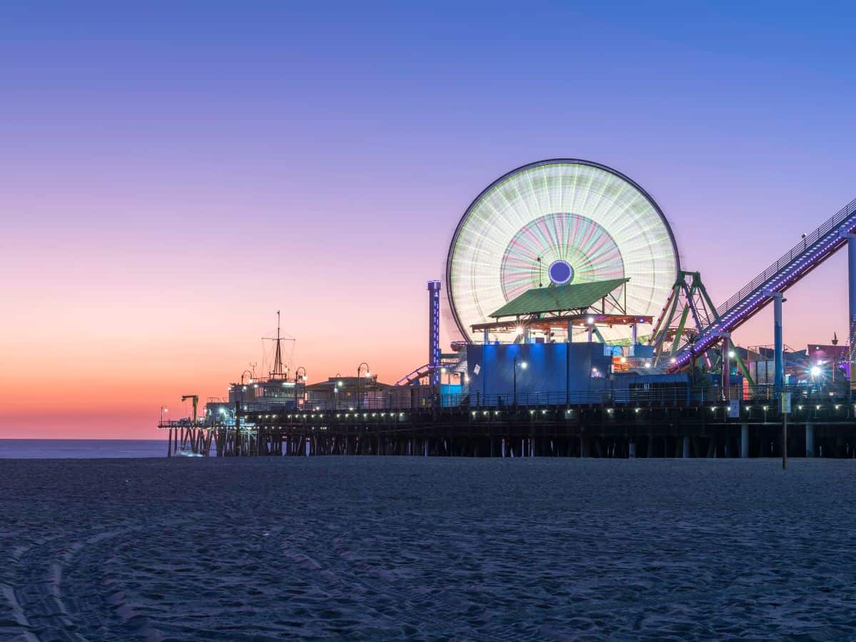 A serene evening shot of Santa Monica Pier with its vibrant Ferris wheel illuminated in neon colors, contrasting with the deep hues of the sunset and the dark beach below.