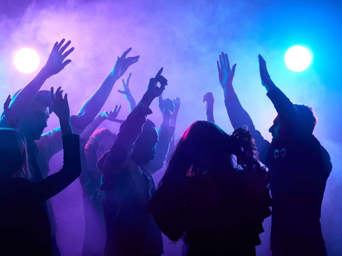 A vibrant club scene with multiple people dancing energetically, their silhouettes illuminated by purple and blue lights amidst a hazy, foggy backdrop.
