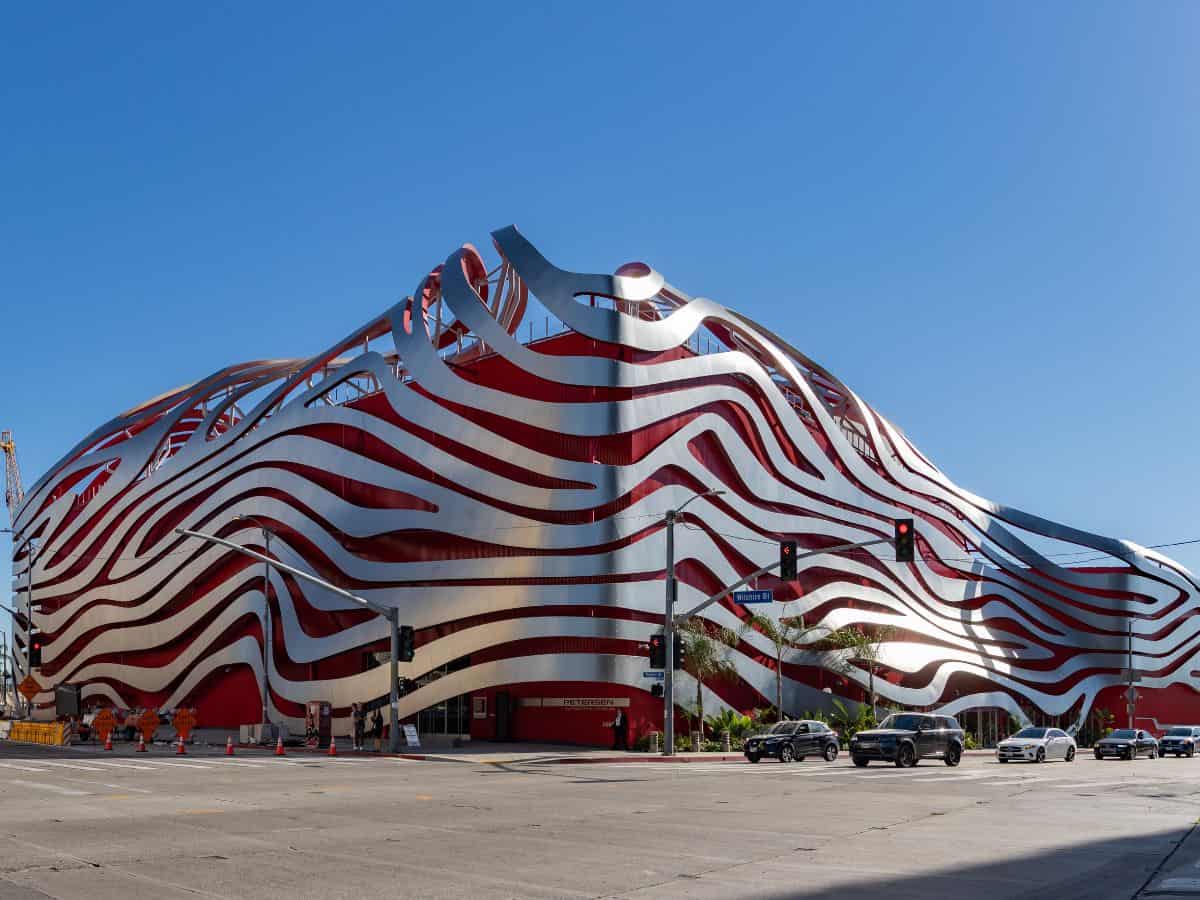 The exterior of the Petersen Automotive Museum in Los Angeles, characterized by its wavy red and silver stripes that ripple across the building's facade, creating a visually striking pattern. The design is modern and artistic, mirroring the innovation of automotive design showcased within the museum.