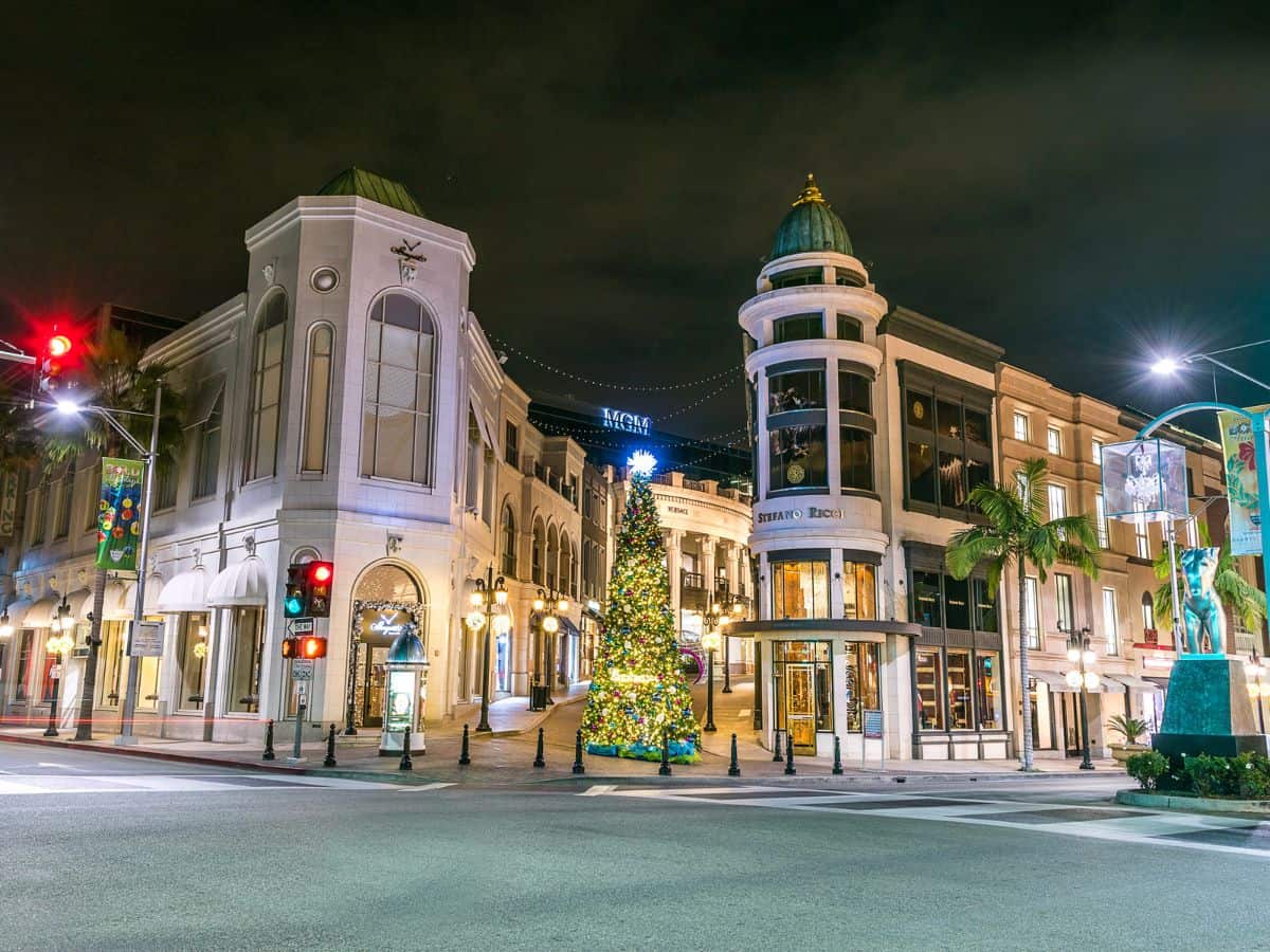 A nighttime scene of Rodeo Drive in Beverly Hills, beautifully decorated with Christmas lights and lavish holiday decorations. The street is lined with high-end boutiques and stores, under a clear night sky.