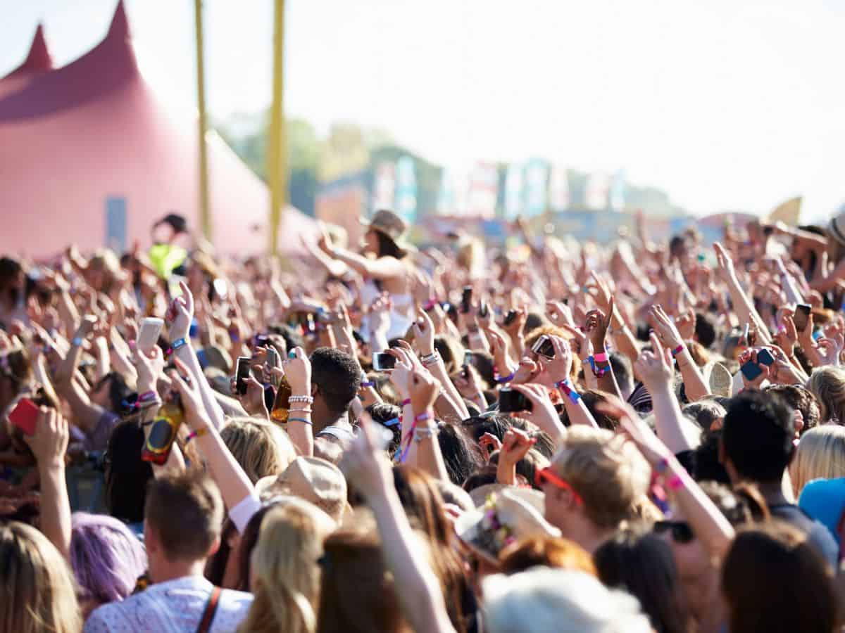 A vibrant and bustling scene at an outdoor music festival, showing a large crowd of festival-goers enjoying the event. People are raising their hands, dancing, and using their smartphones to capture the moment.