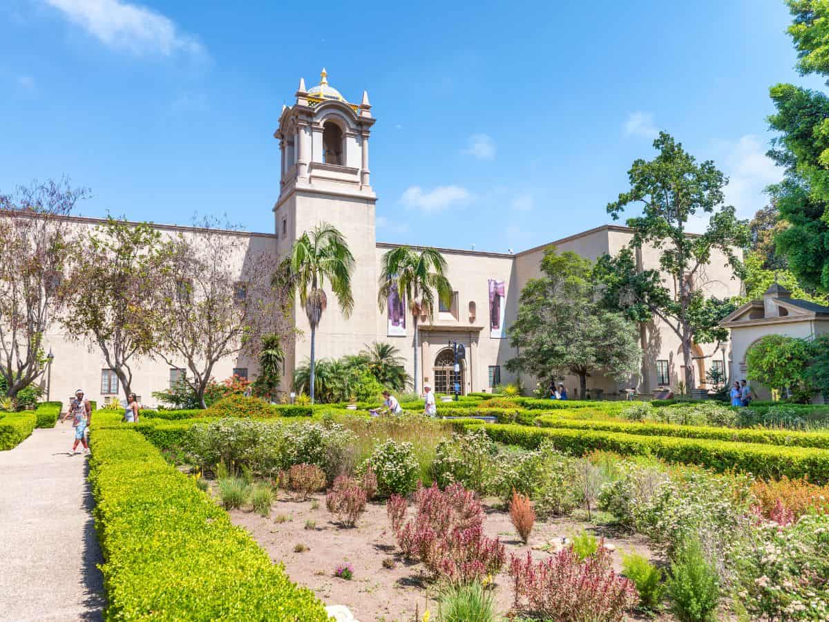 he lush gardens and historical architecture of Balboa Park, a highlight among things to do in San Diego, during Museum Month. The park's well-maintained garden beds, filled with colorful plants, are framed by elegant Spanish-Renaissance-style buildings under a clear blue sky.
