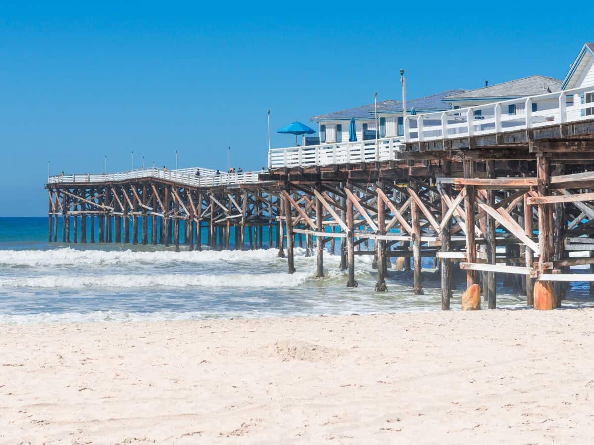 The long wooden pier at Pacific Beach, a classic attraction among things to do in San Diego, stretching out into the blue ocean. The white sandy beach is bathed in sunlight, and the waves gently roll towards the shore, inviting beachgoers to relax and enjoy the coastal vibe.