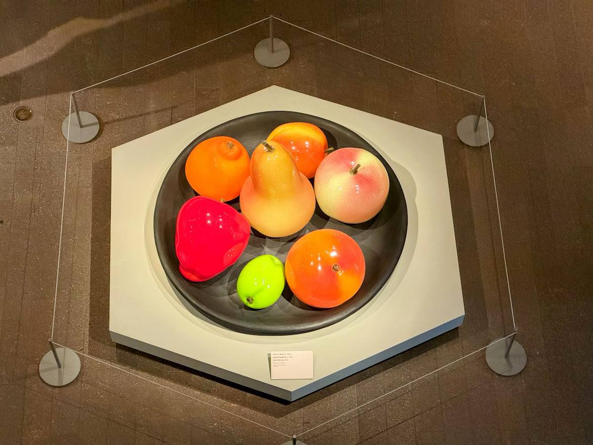 A colorful fruit bowl art installation featuring oversized, glossy sculptures of various fruits including an apple, pear, orange, and bell pepper, displayed in a modern gallery setting with protective barriers around the artwork.