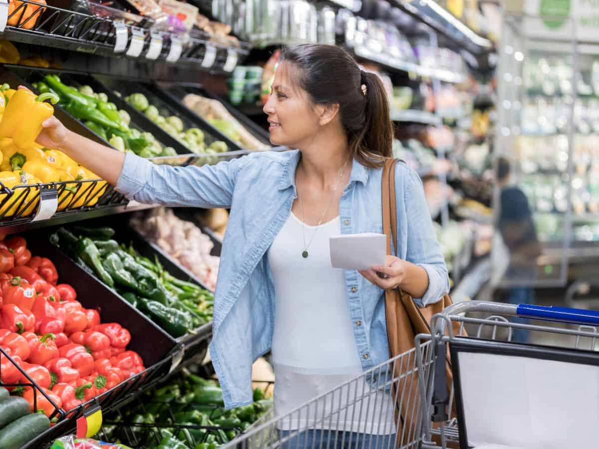 A woman shopping in a grocery store, reaching for fresh produce with a list in hand. The high demand and limited supply of groceries contribute to California's high cost of living, explaining why it is so expensive.