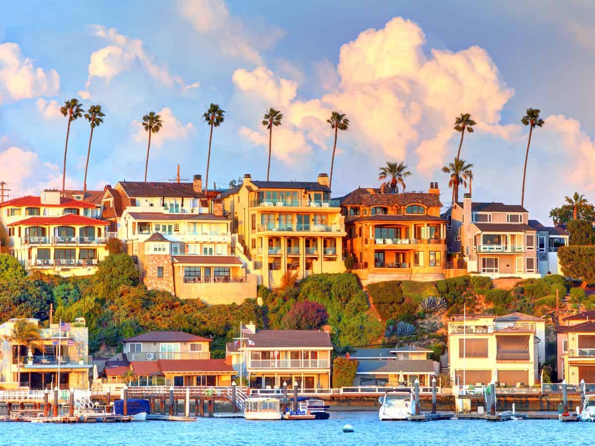A hillside view of upscale houses overlooking the ocean at sunset. The high demand and low supply of housing lead to increased property values, explaining why California is so expensive.