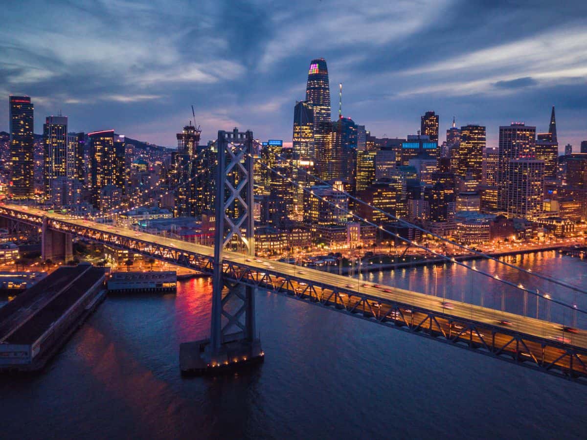 An aerial view of the San Francisco skyline at dusk, showcasing the brightly lit cityscape and the Bay Bridge crossing the water. The scene captures California's urban density and infrastructure, which partially explains why California is so expensive due to high living costs, including elevated demand for electricity in densely populated areas.