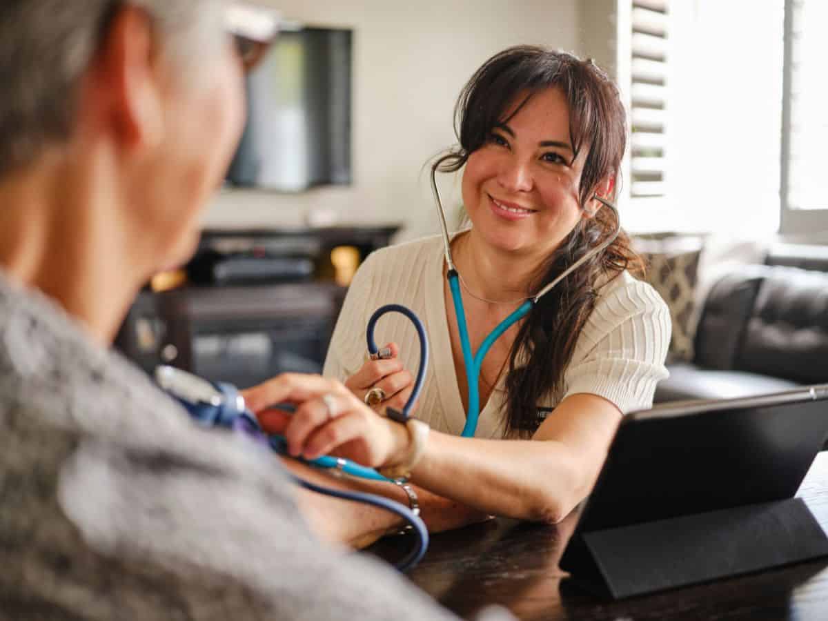 A smiling healthcare professional checking the blood pressure of an elderly patient in a cozy living room. The high cost of healthcare services in California is another factor that explains why California is so expensive.