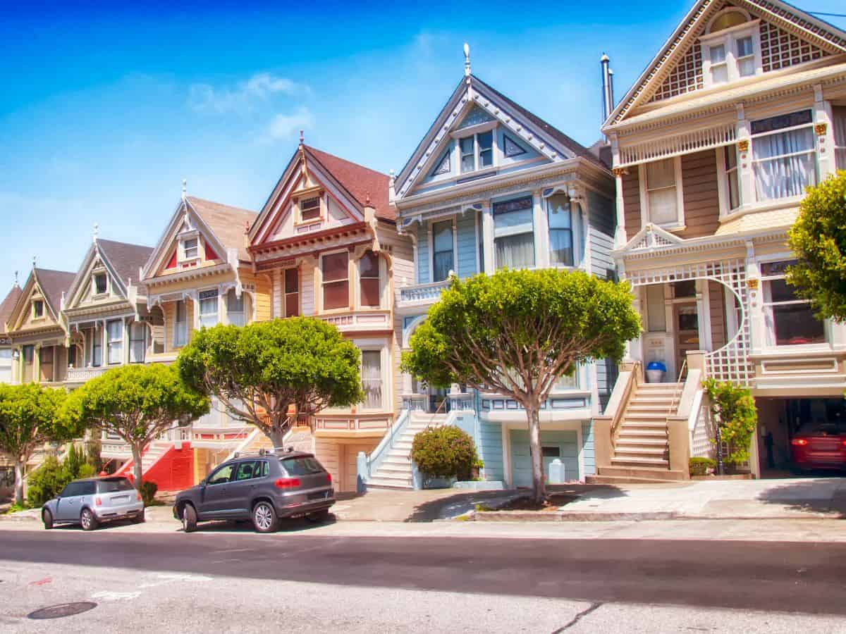 A row of iconic, colorful Victorian houses on a sunny day. The high cost of homes in California, influenced by various economic factors, is a major reason why California is so expensive.