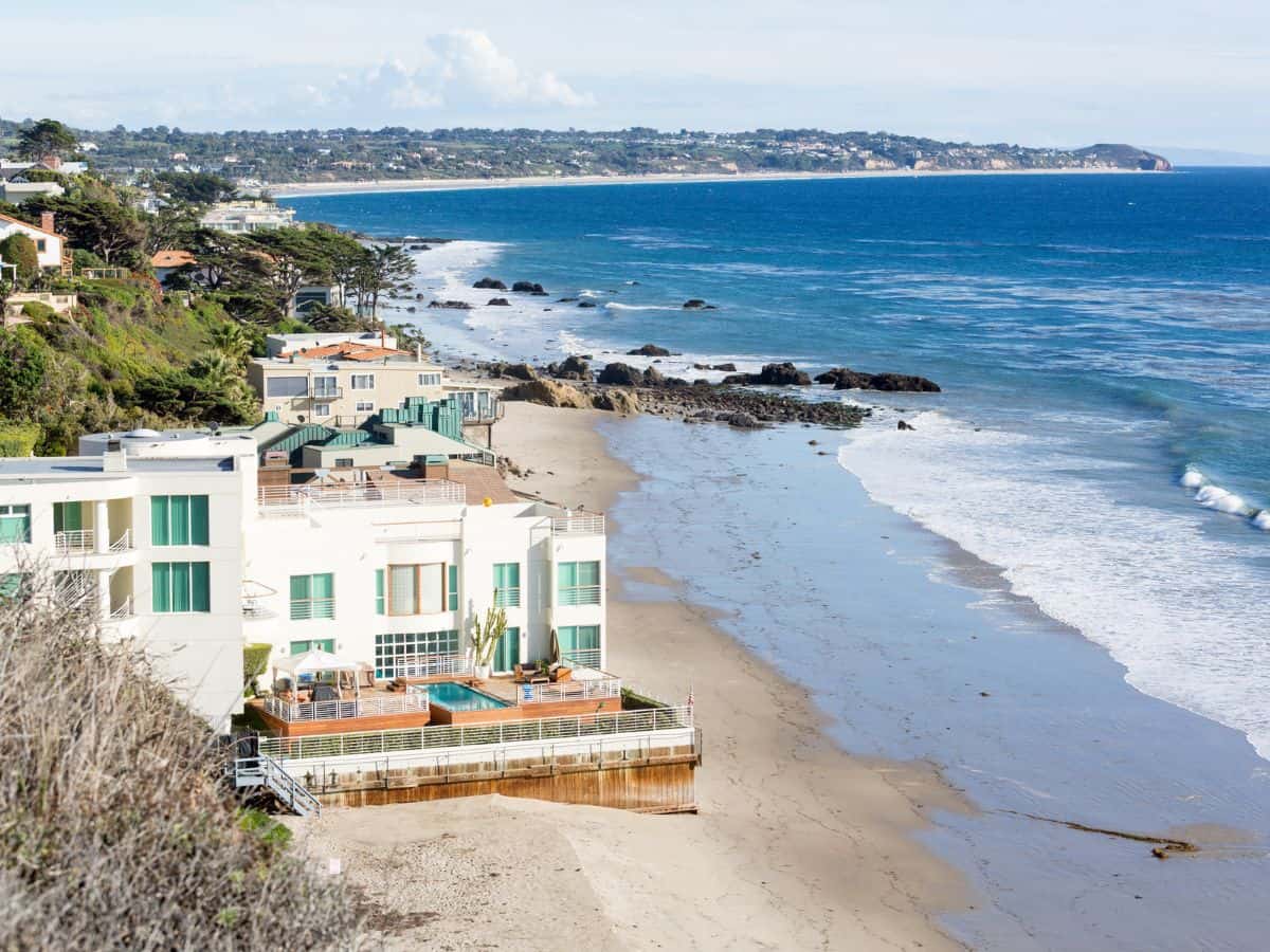 A scenic coastal view with luxury homes along a sunny California beach. High property taxes in these areas are a significant factor in why California is so expensive.