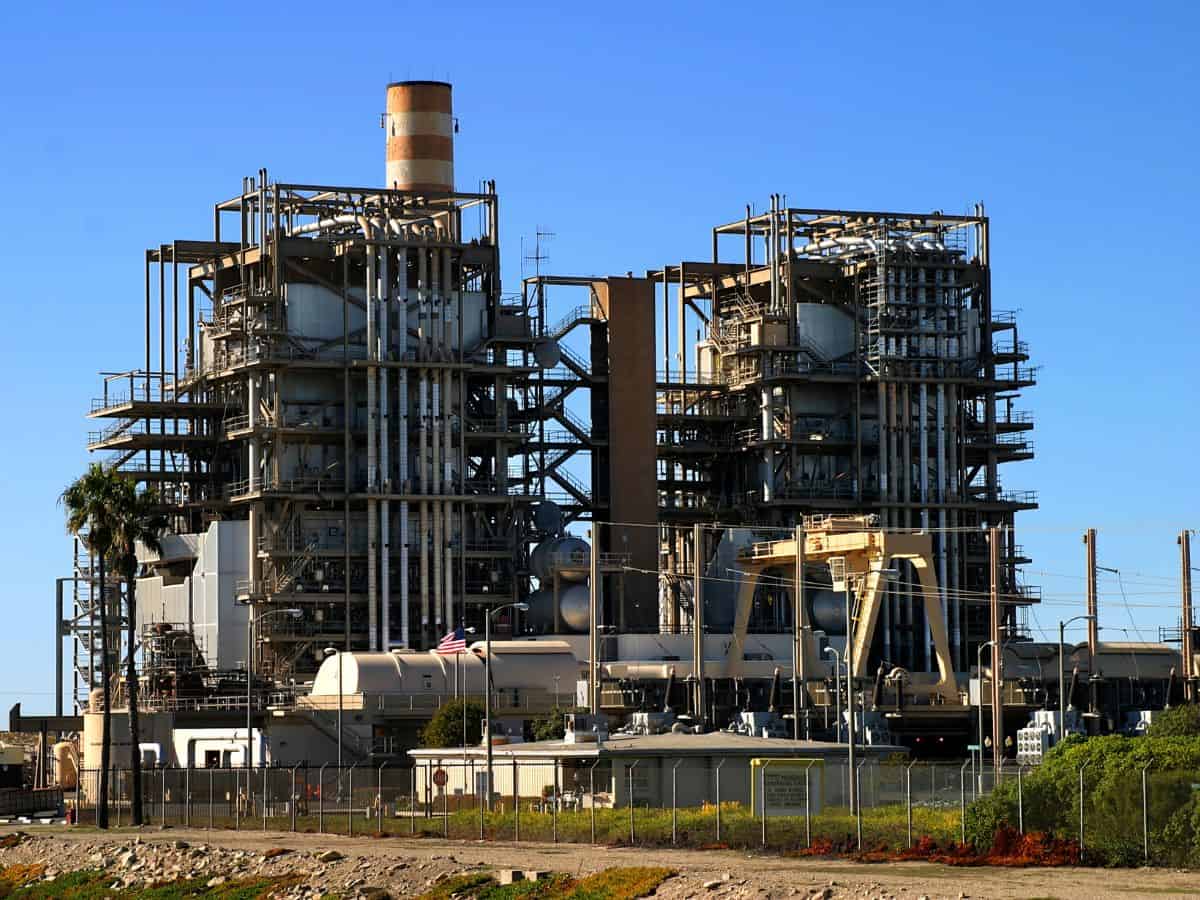 An industrial power plant with tall stacks and intricate piping under a clear blue sky. High taxes on energy production and usage in California lead to higher utility bills, one of the reasons why California is so expensive.