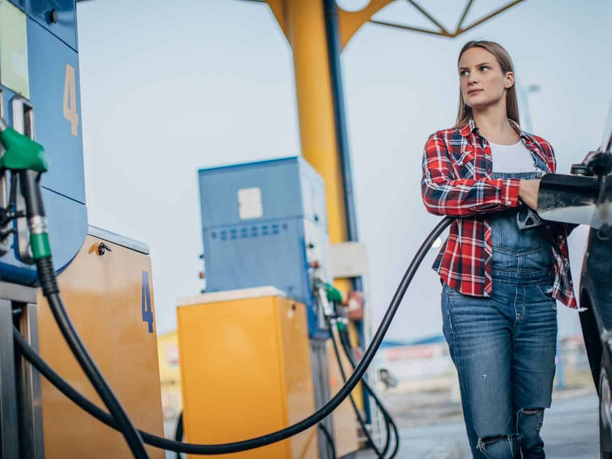 A young woman in a plaid shirt is refueling her car at a gas station. The pump displays high numbers, hinting at the elevated gas prices in California. Out-of-state transportation costs contribute significantly to why California is so expensive.
