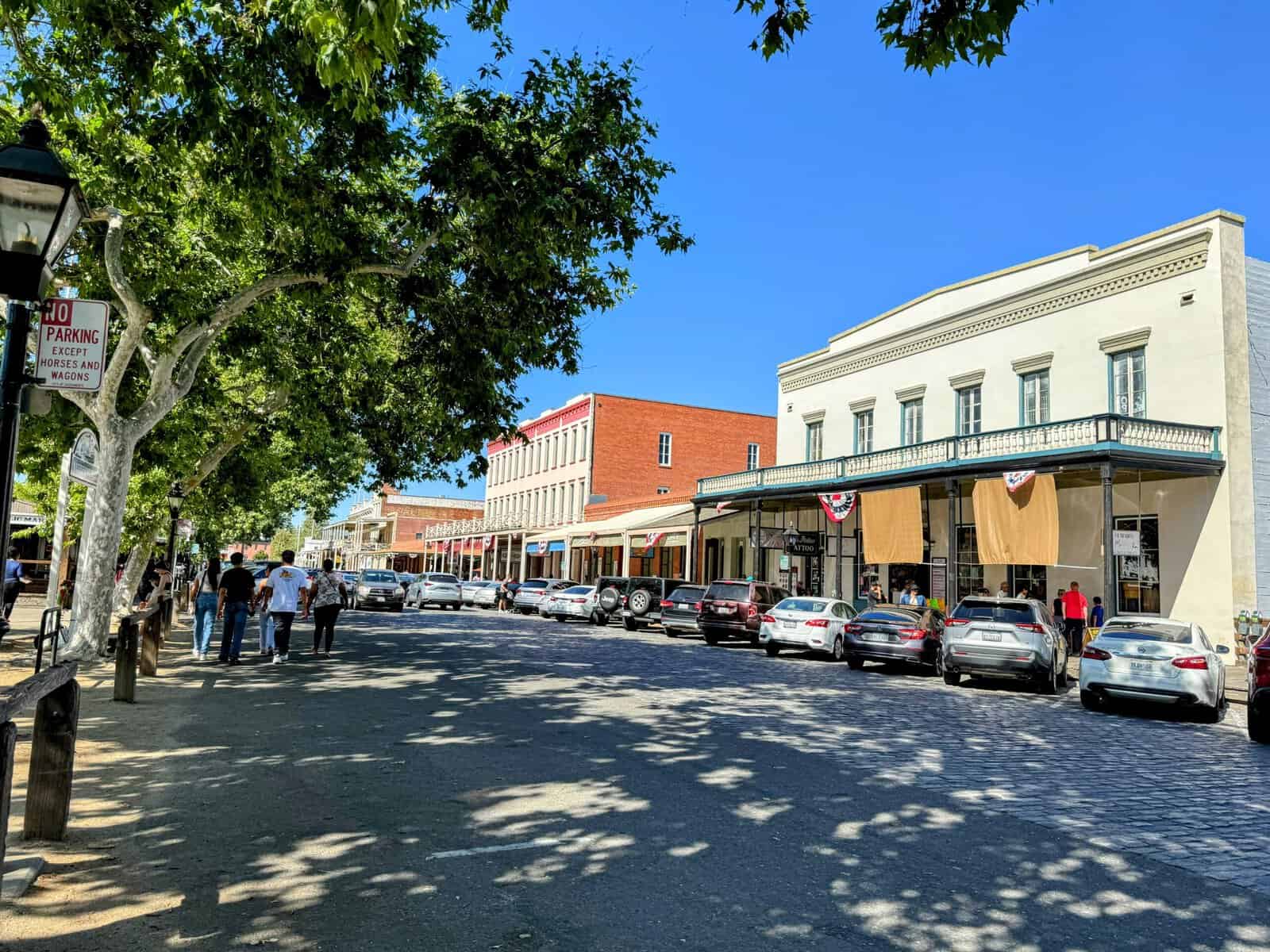 This photo shows a bustling street in Old Sacramento, with historic buildings, parked cars, and people walking under large shady trees. The cobblestone street and a "No Parking Except Horses and Wagons" sign add a charming, nostalgic feel to the scene, all under a bright blue sky.