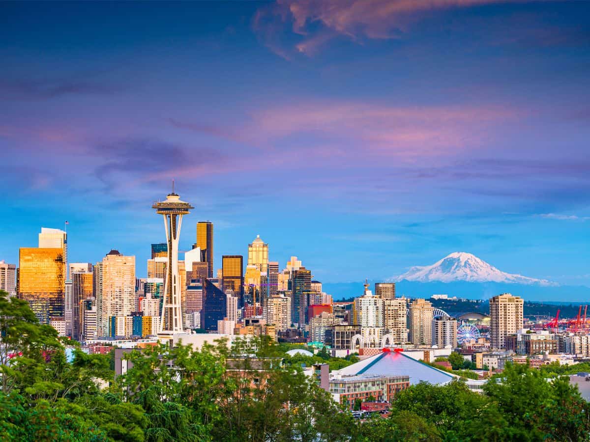 The Seattle skyline glows under a pastel sunset, with the Space Needle standing tall among modern high-rises. Mount Rainier, snow-capped and majestic, rises in the distance behind the city. Lush greenery in the foreground adds to the vibrant urban-meets-nature scene.