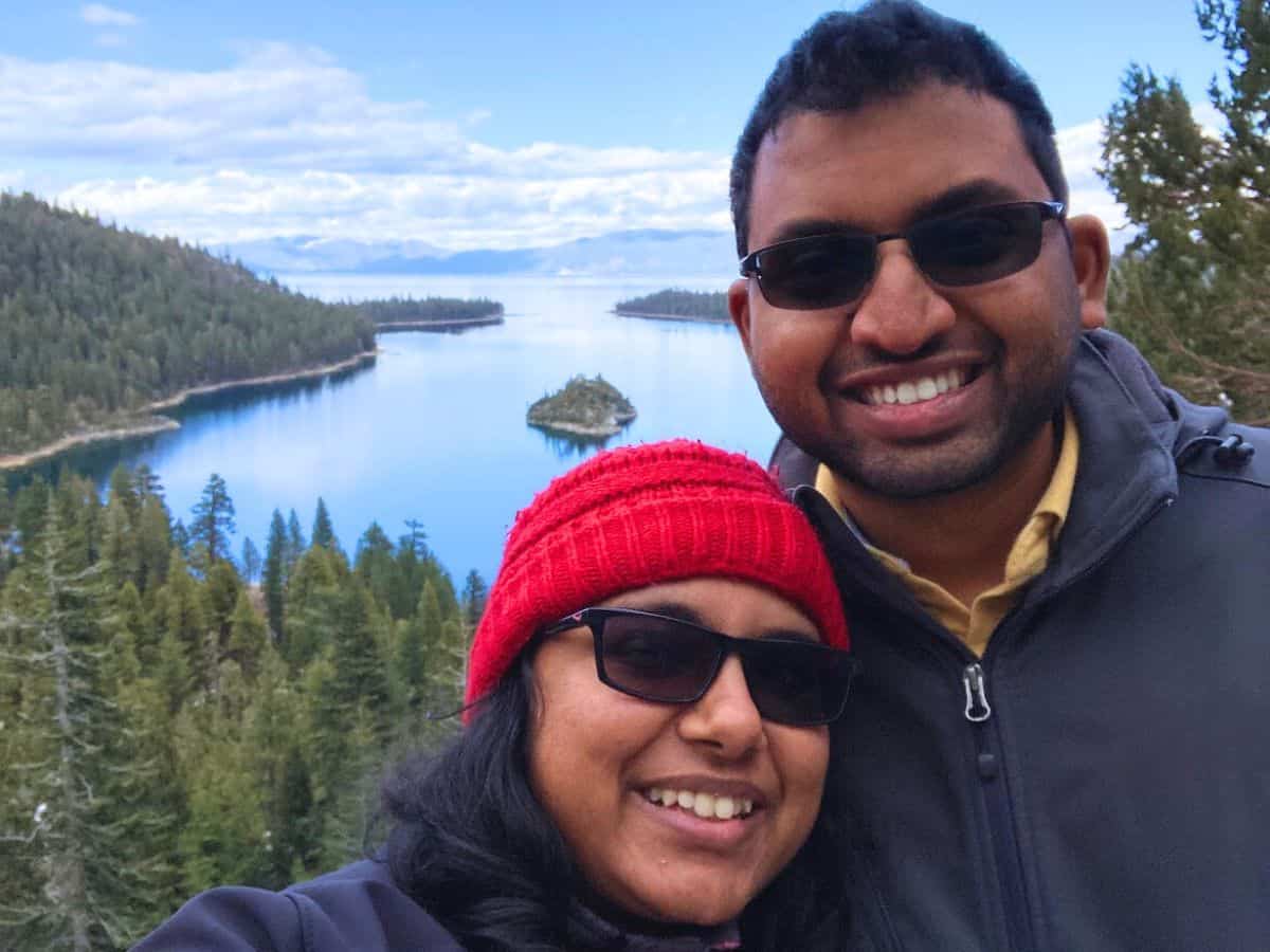 A smiling couple poses in front of the serene waters of South Lake Tahoe, with lush green pine trees and a small island in the background, highlighting the peaceful and scenic beauty of the area’s beaches.