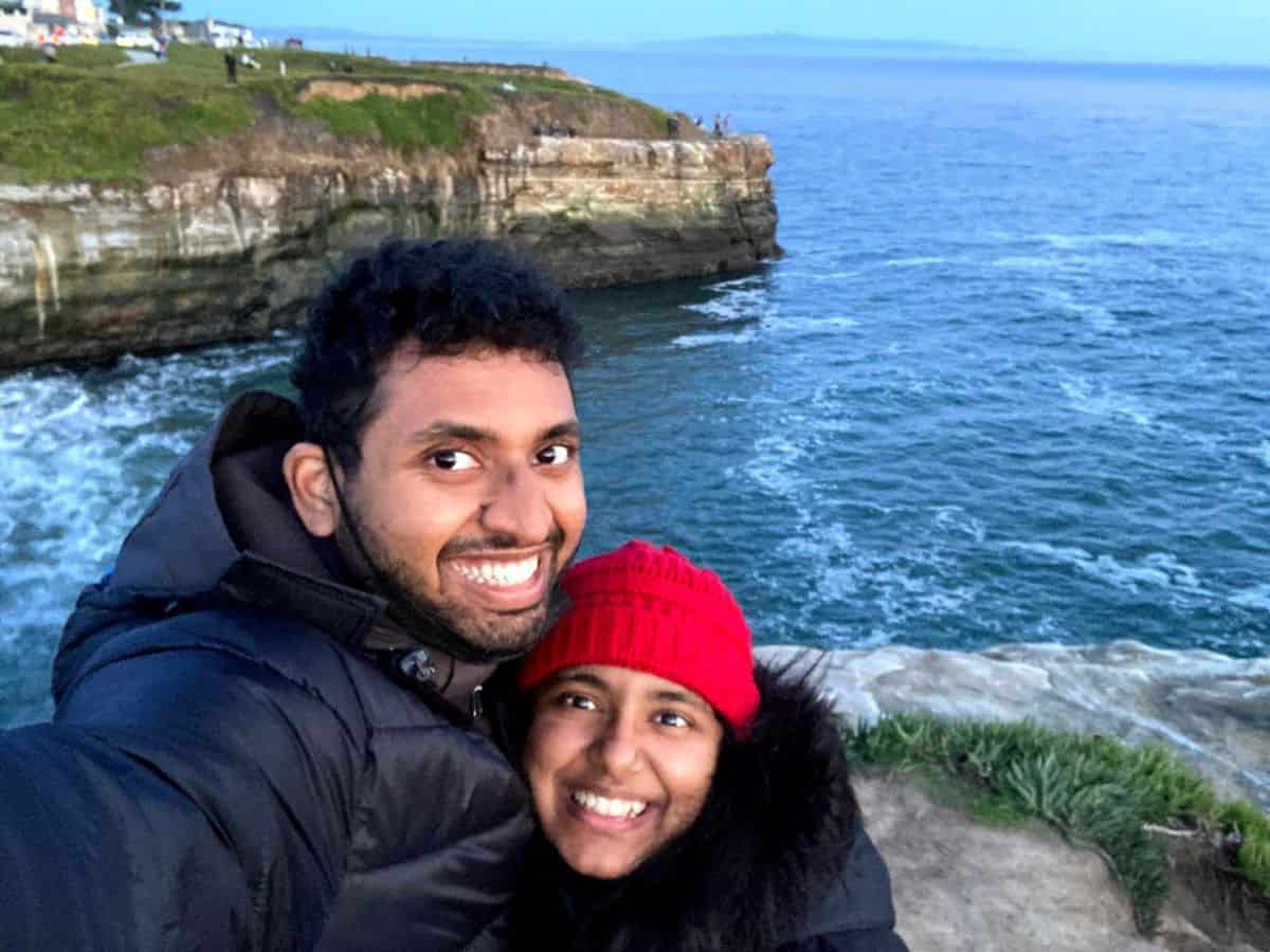 A happy couple takes a selfie near the edge of a scenic cliff overlooking the ocean at sunset, with the rugged coastline and waves creating a stunning backdrop, capturing the charm of Santa Cruz beaches.