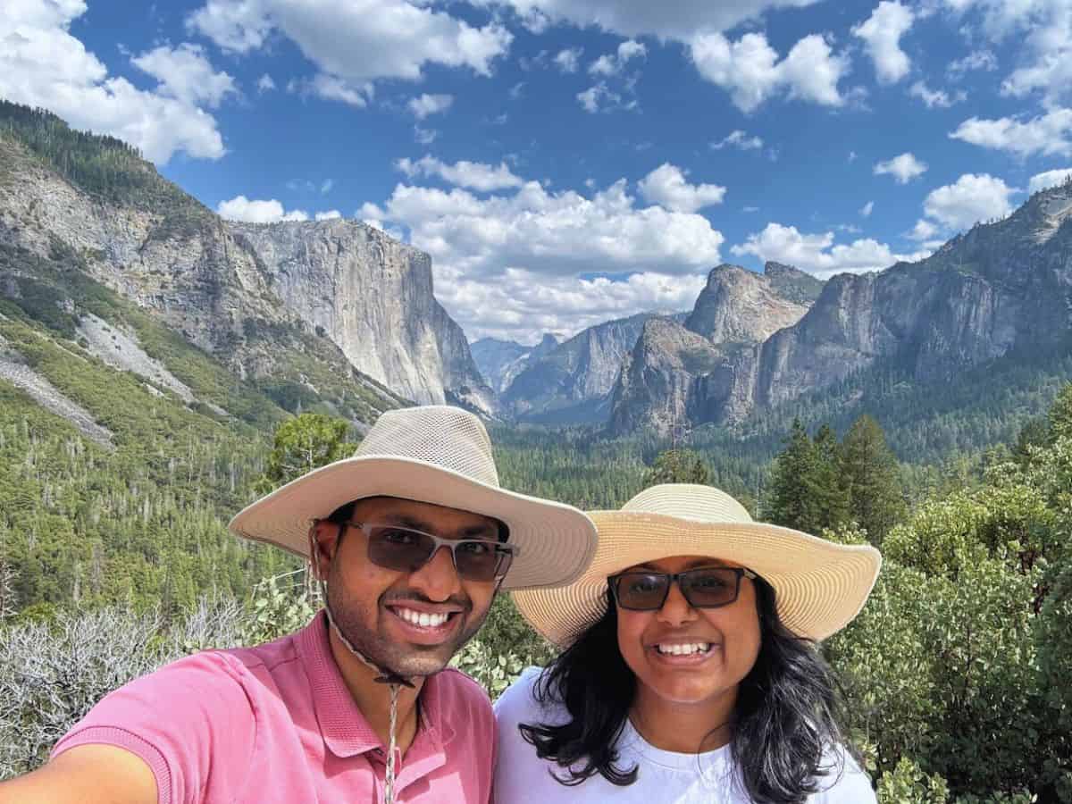 A smiling couple takes a selfie while enjoying a stunning view of Yosemite National Park's iconic granite cliffs and lush forests under a bright blue sky with scattered clouds, highlighting one of the park's breathtaking viewpoints.