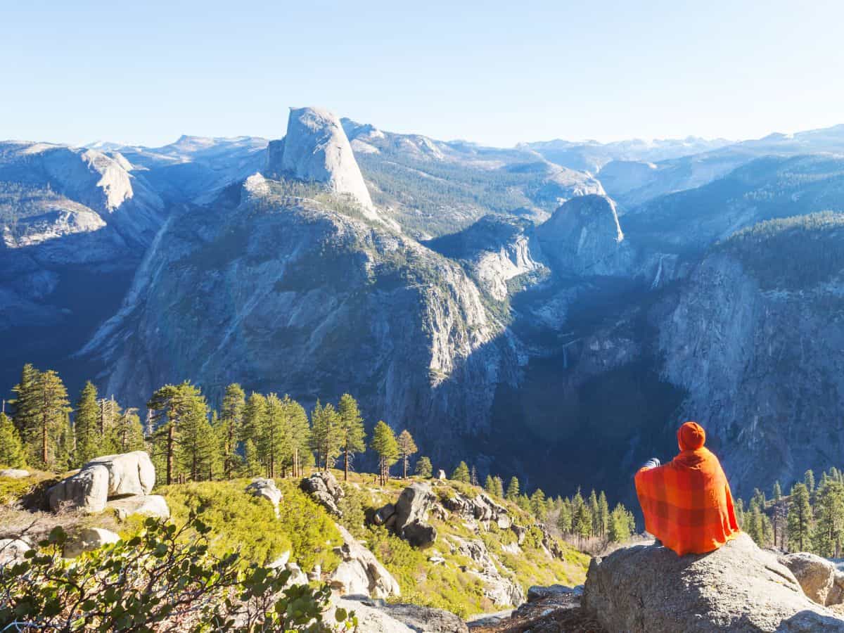 A traveler wrapped in an orange blanket sits on a rock, gazing out at the awe-inspiring view of Yosemite's Half Dome and surrounding valleys under a clear blue sky. This serene moment captures the majestic beauty and tranquility of one of the must-visit spots on the Death Valley to Yosemite road trip.