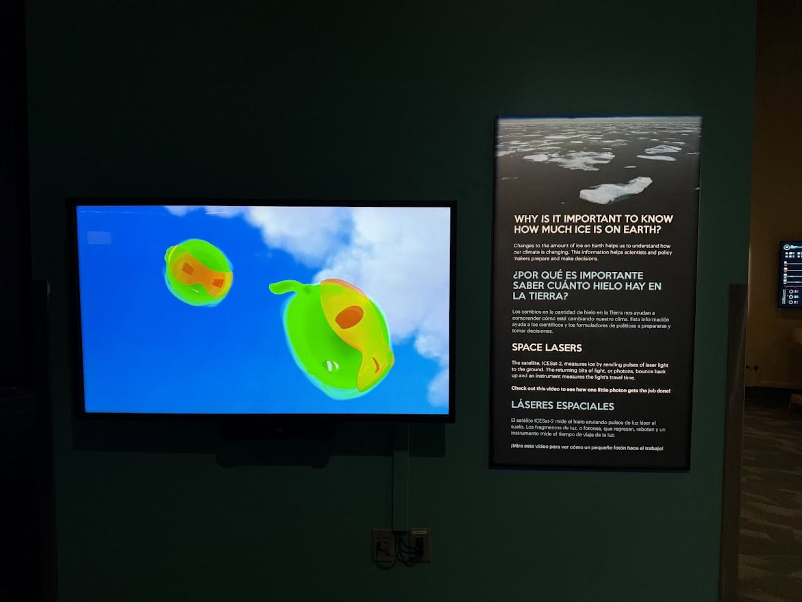 An educational exhibit showing a screen with colorful photon graphics and a sign explaining the role of the ICESat-2 satellite in measuring Earth's ice levels, demonstrating the importance of this data in understanding climate change.
