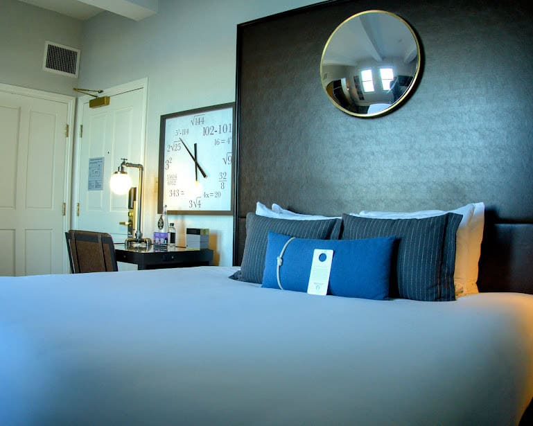 A cozy guest room at the Harbor Court Hotel in San Francisco, showcasing a plush bed with blue accent pillows, a quirky math-themed wall clock, and stylish decor in a minimalist design.