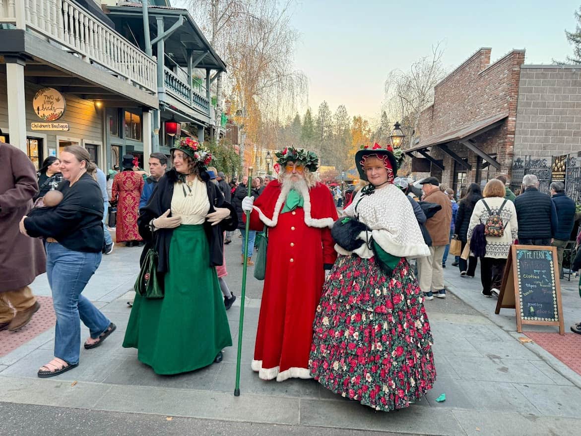 Father Christmas posing with two Victorian-dressed carolers at the Nevada City Victorian Christmas. The trio stands in front of decorated shop windows, exuding holiday spirit.
