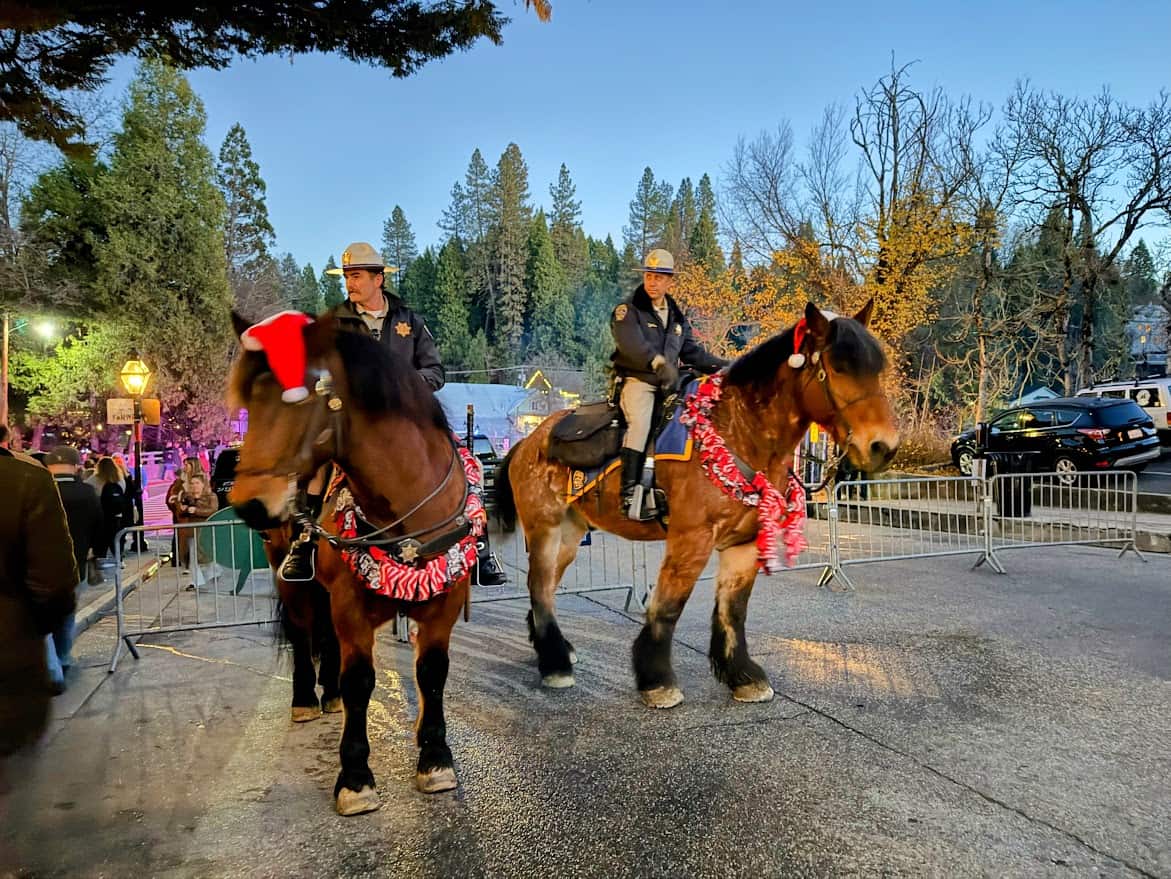 Two officers on horseback dressed in festive holiday gear, including red bows and Santa hats, at the Nevada City Victorian Christmas. The horses stand near a barricaded area with trees and twinkling lights in the background.