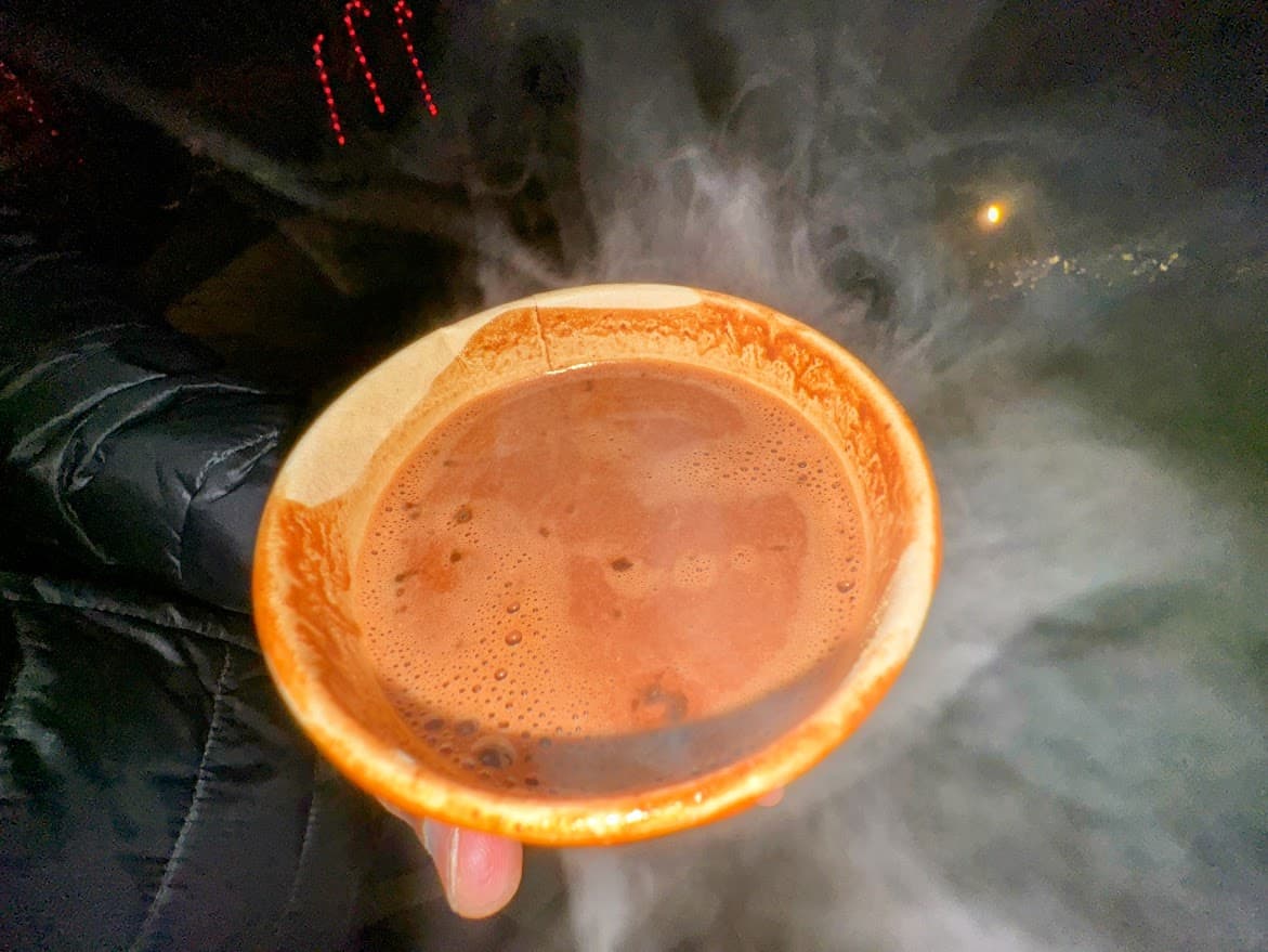 A steaming cup of rich hot chocolate from Choquiero Cacao Cafe at the Nevada City Victorian Christmas. The warm drink is served in a paper cup, with a misty evening backdrop creating a cozy holiday atmosphere.