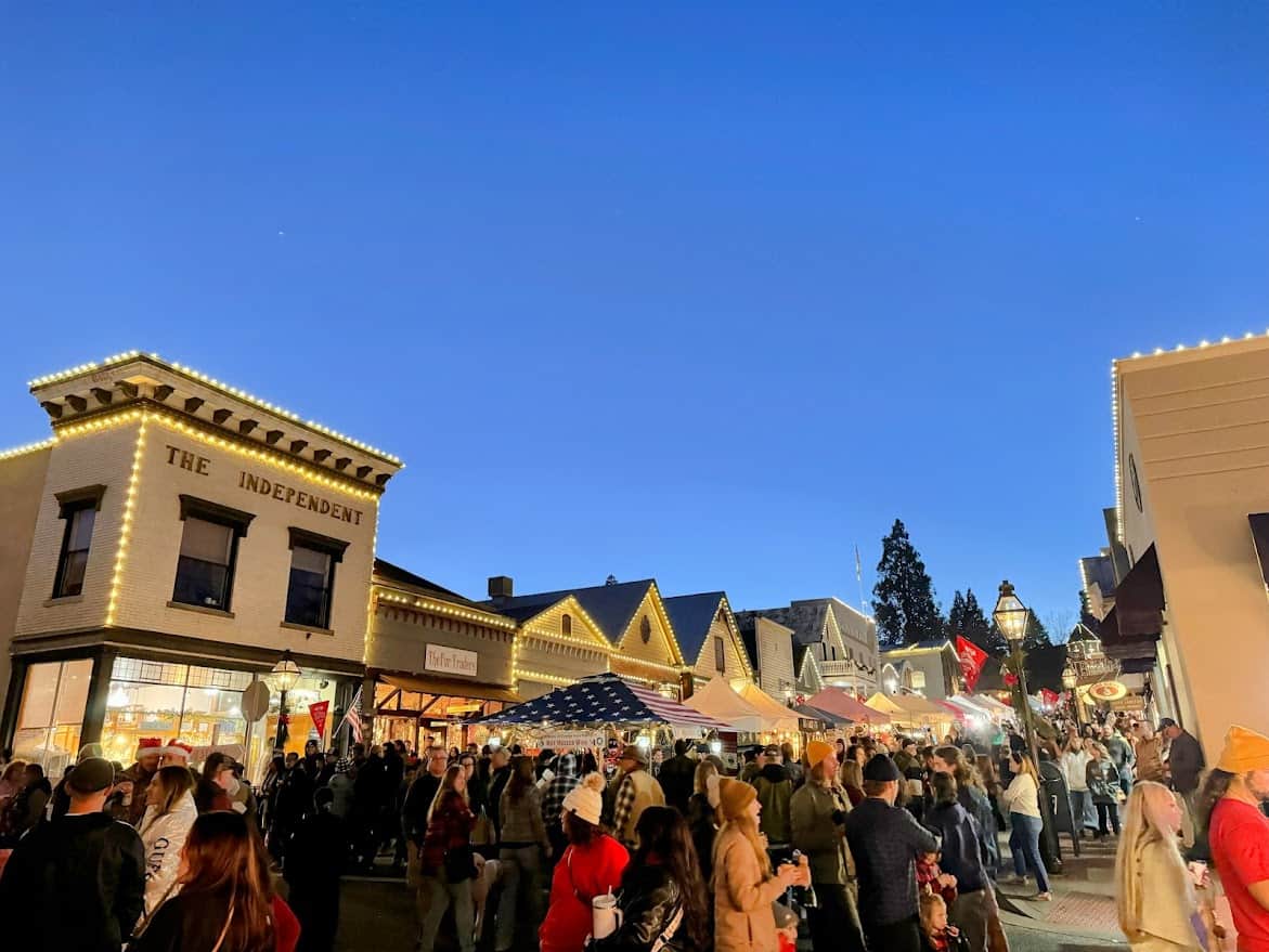 A view of Nevada City's Victorian Christmas at night, showcasing brightly lit buildings, festive market stalls, and a bustling crowd enjoying the holiday ambiance under a clear, dark sky.