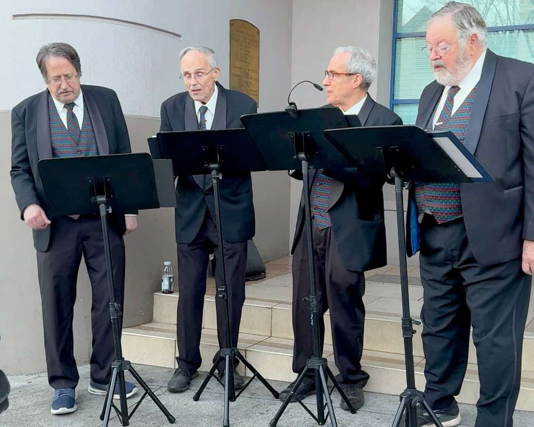 A group of performers dressed in black suits and festive vests singing outdoors at the Nevada City Victorian Christmas. They are standing with microphones in front of a building, providing live entertainment for attendees.