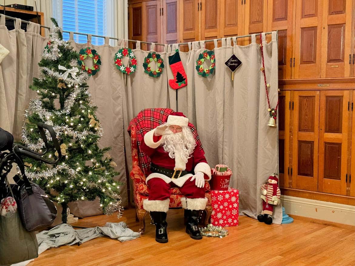 Santa Claus seated in a cozy holiday setup at the Nevada City Victorian Christmas, surrounded by a decorated Christmas tree, wreaths, and wrapped presents.