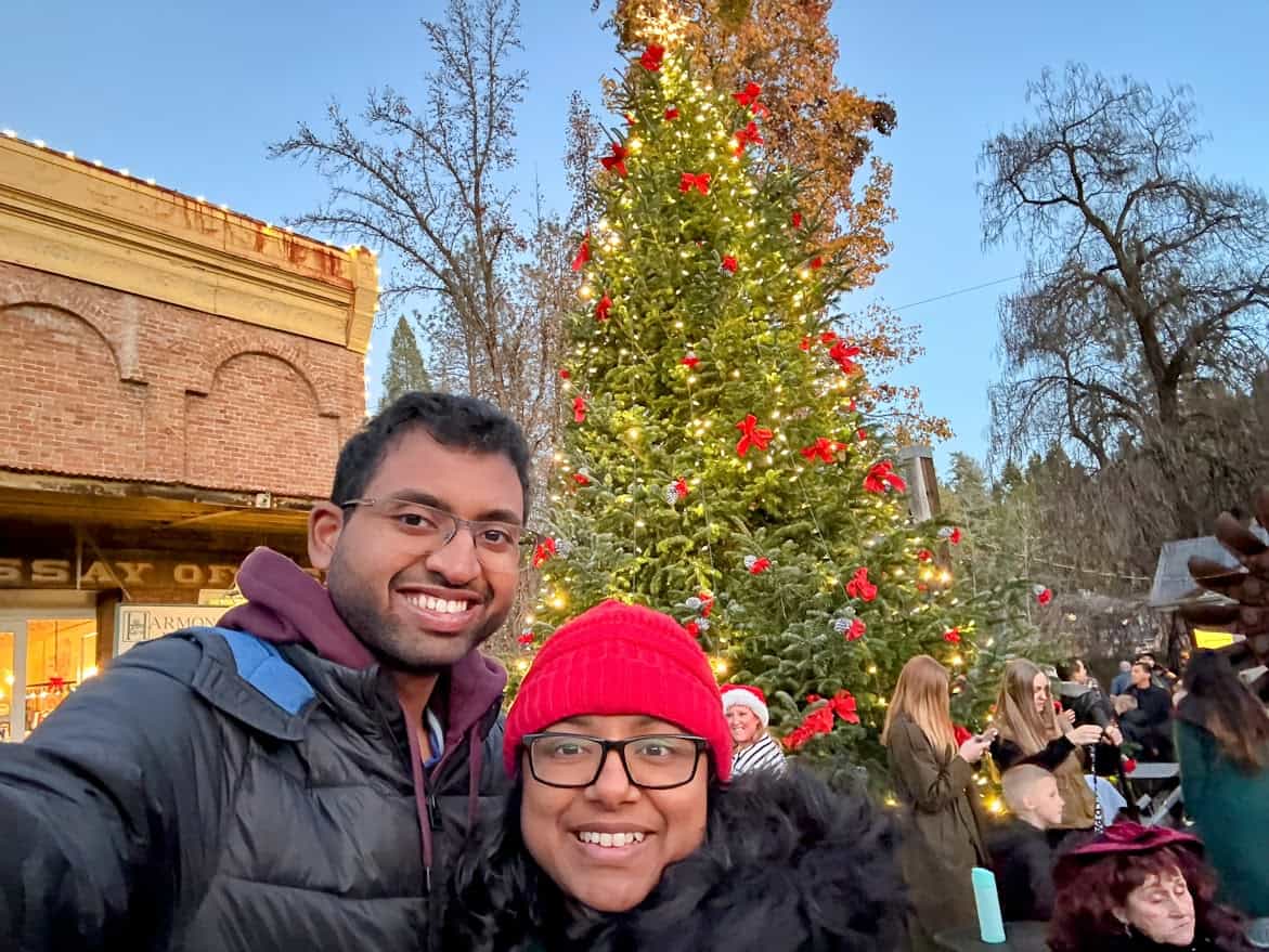 Shreeyeh and Kiran take a selfie in front of the 25-foot Christmas tree at the Nevada City Victorian Christmas. The tree is adorned with red bows and glowing lights, surrounded by festive visitors enjoying the event.
