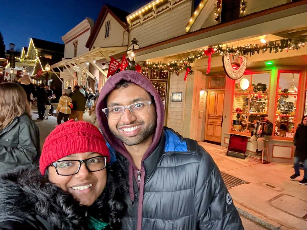 A couple bundled up in winter coats and hats, smiling in front of beautifully lit Victorian-era shops at the Nevada City Victorian Christmas. The glowing storefronts and festive garlands create a charming holiday backdrop.