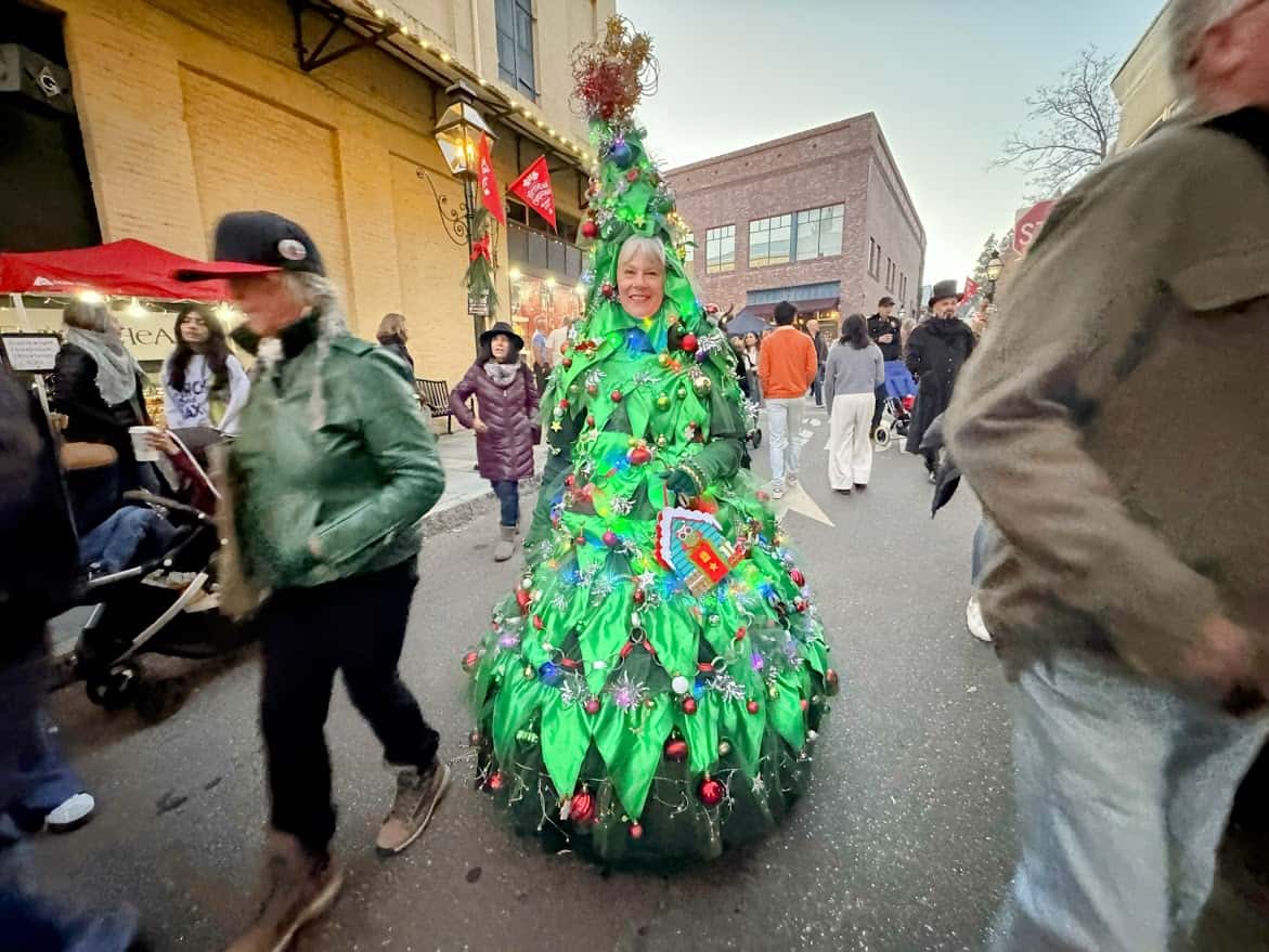 A cheerful participant dressed as a walking Christmas tree at the Nevada City Victorian Christmas. The costume is vibrant green, adorned with ornaments, colorful lights, and festive garlands.