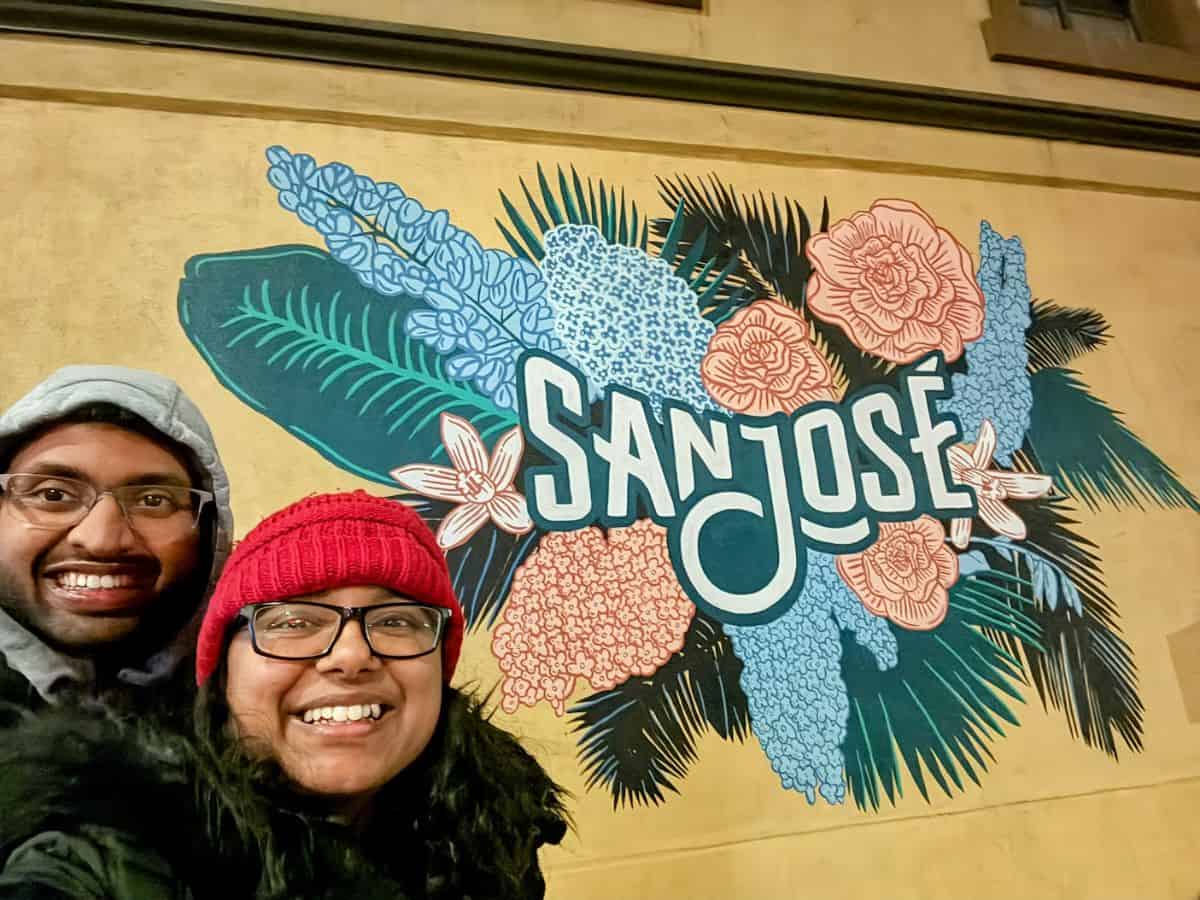 A cheerful photo of two people smiling in front of a vibrant mural featuring the words "San Jose" surrounded by colorful floral and leafy designs. This image captures the lively and welcoming vibe of exploring the 10 Vegan Spots in San Jose.