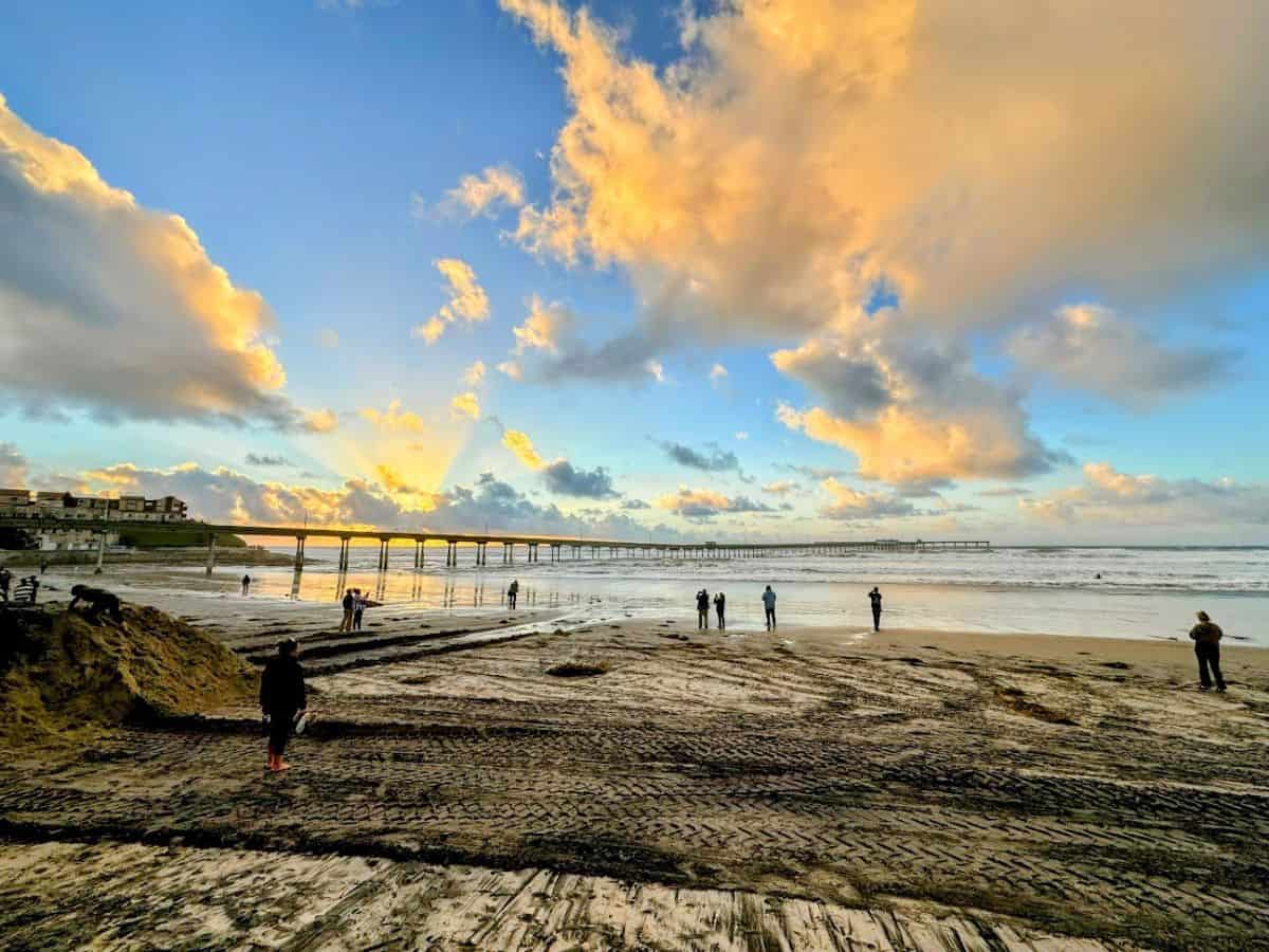 A scenic sunset at Ocean Beach Pier in San Diego, with golden clouds reflecting over the wet sand as people stroll along the shoreline.