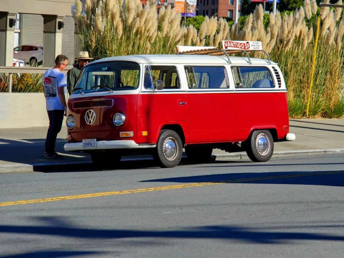 A vintage red and white Volkswagen van parked on a street, with two men standing nearby engaged in conversation. The van has a sign on top reading "Vantigo SF," hinting at local tour services. The scene captures the charm of classic vehicles in San Francisco while reflecting on the high costs associated with car maintenance and ownership, contributing to the broader question, Why Is San Francisco So Expensive.